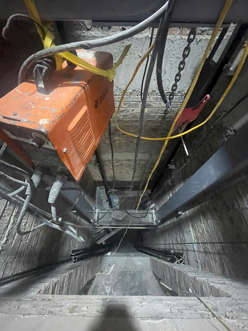 Hố thang máy đang thi công với máy tời màu cam và hệ thống cáp điện, ray dẫn hướng